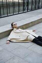 Androgynous man wearing poncho and sleeping on concrete street in city - EKHF00084