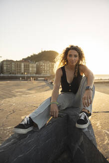 Young woman with curly hair sitting on promenade - MTBF01279