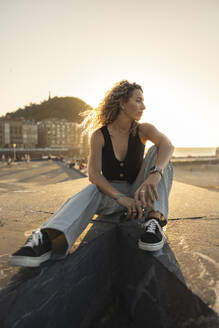 Contemplative woman with curly hair sitting on promenade - MTBF01280