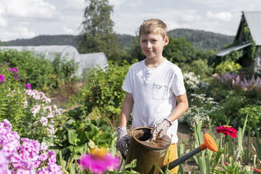 Lächelnder Junge steht mit Gießkanne im Garten an einem sonnigen Tag - VBUF00374