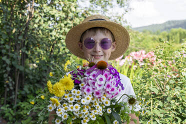 Glücklicher Junge mit Hut, der verschiedene Blumen im Garten hält - VBUF00385