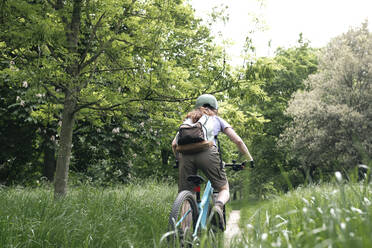 Young woman with backpack riding bicycle on grass in forest - AMWF01663