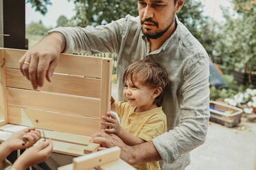 Vater hilft glücklichen Söhnen bei der Herstellung einer Holzkiste im Hinterhof - ANAF02024