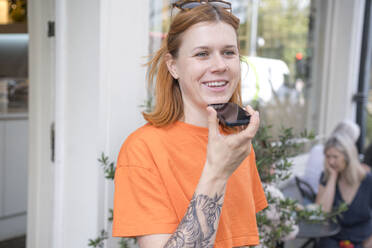 Lächelnde Frau, die eine Sprachnachricht über ein Mobiltelefon in einem Straßencafé sendet - AMWF01689
