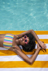 Glückliche junge Frau, die auf einem gestreiften Handtuch am Swimmingpool liegt - VRAF00133