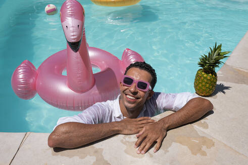 Glücklicher Mann lehnt mit Ananas und aufblasbarem Schwimmring am Pool - VRAF00136