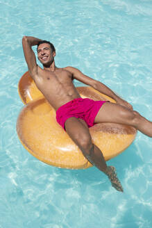 Glücklicher junger Mann sitzt auf einem aufblasbaren Schwimmer im Pool - VRAF00137