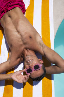 Lächelnder Mann mit Sonnenbrille entspannt sich auf einem Handtuch am Pool - VRAF00147