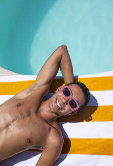 Lächelnder Mann mit Sonnenbrille entspannt sich auf einem Handtuch am Pool - VRAF00148