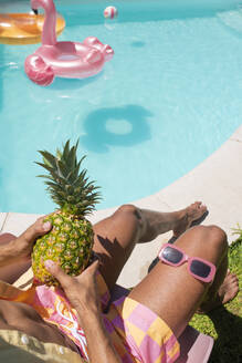 Junger Mann sitzt mit Ananas und Sonnenbrille am Pool - VRAF00150