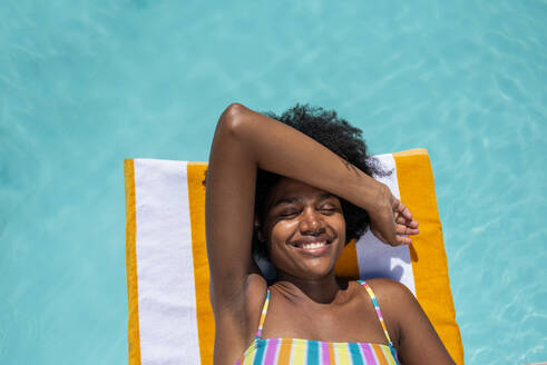 Lächelnde junge Frau, die mit geschlossenen Augen im Schwimmbad auf einem gestreiften Handtuch liegt - VRAF00159