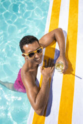 Lächelnder junger Mann mit Getränk, der sich auf ein Handtuch am Pool stützt - VRAF00176