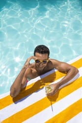 Glücklicher junger Mann mit Getränk, der sich auf ein Handtuch am Pool stützt - VRAF00177