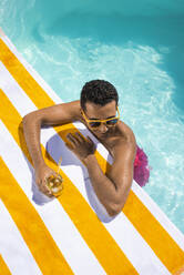 Lächelnder Mann mit Getränk, der sich auf ein Handtuch am Pool stützt - VRAF00179