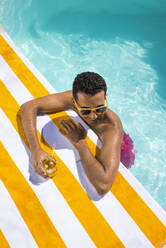 Smiling man with drink leaning on towel at poolside - VRAF00179