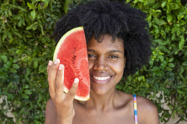 Lächelnde junge Frau mit Scheibe Wassermelone vor Pflanzen - VRAF00180