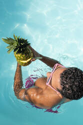Man holding pineapple in swimming pool on sunny day - VRAF00190