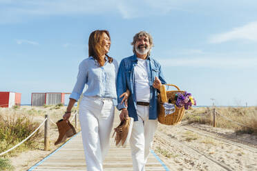 Glückliche ältere Frau und Mann mit Picknickkorb und Schuhen beim Spaziergang am Strand - OIPF03447