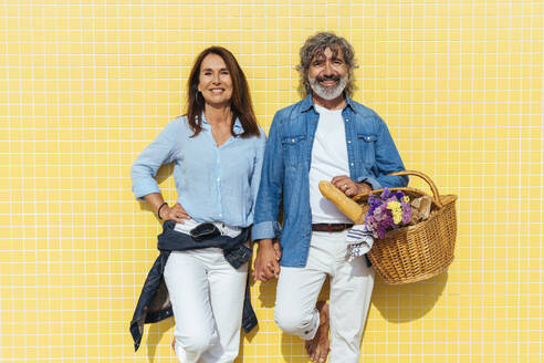 Happy senior couple with picnic basket standing in front of yellow wall - OIPF03449