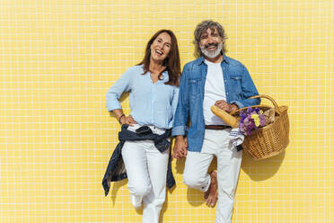 Fröhlicher älterer Mann und Frau mit Picknickkorb vor einer gelben Wand - OIPF03450