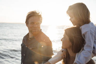 Smiling father looking at mother carrying daughter on shoulders at beach - JOSEF20633