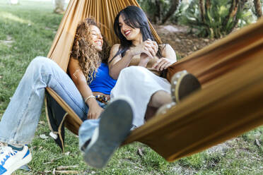 Smiling lesbian couple holding hands and lying in hammock - PBTF00231