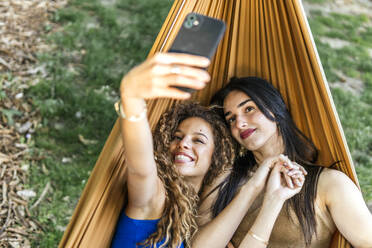 Smiling young girlfriends taking selfie on mobile phone in hammock - PBTF00235