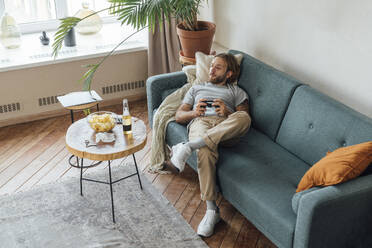 Man with game controller lying on sofa at home - VPIF08470