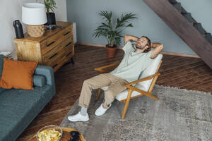 Man with hands behind head relaxing on armchair at home - VPIF08483