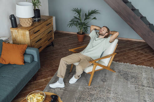 Man with hands behind head relaxing on armchair at home - VPIF08483