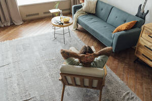 Mann mit Händen hinter dem Kopf entspannt auf Sessel im Wohnzimmer zu Hause - VPIF08484