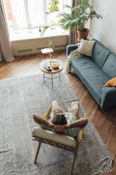 Young man with hands behind head relaxing on armchair at home - VPIF08485