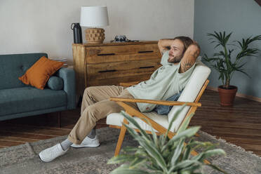 Thoughtful young man with hands behind head relaxing on armchair at home - VPIF08486