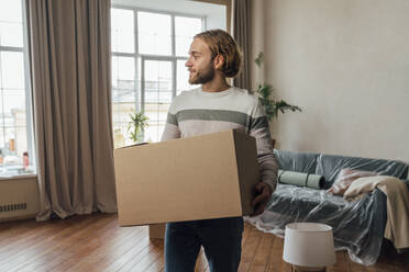 Young man with cardboard box standing in living room - VPIF08521