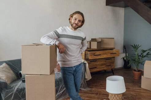 Smiling young man standing by cardboard boxes in living room - VPIF08524