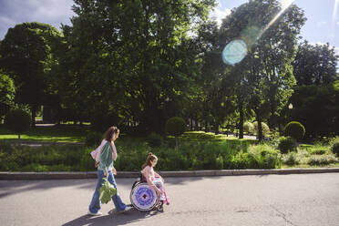 Daughter in wheelchair with mother walking behind at park on summer - EYAF02726