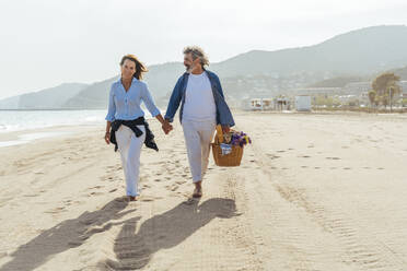 Glücklicher älterer Mann, der einen Korb hält und mit einer Frau am Strand spazieren geht - OIPF03474