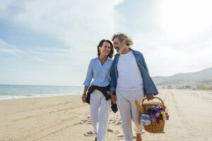 Glückliches Paar hält sich an den Händen und geht mit einem Korb am Strand spazieren - OIPF03475
