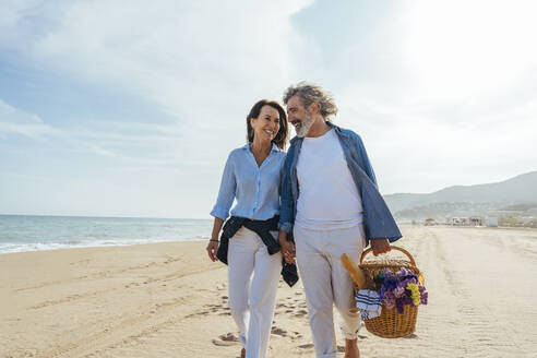 Happy couple holding hands and walking with basket at beach - OIPF03475