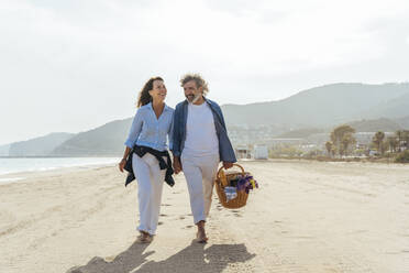 Glückliches Paar, das sich an den Händen hält und mit einem Korb am Strand spazieren geht, an einem sonnigen Tag - OIPF03477