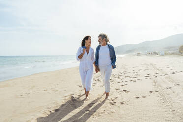 Lächelnder älterer Mann und Frau spazieren am Strand an einem sonnigen Tag - OIPF03486