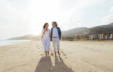 Happy couple holding hands and walking at beach under sky on sunny day - OIPF03492