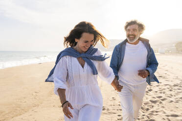 Fröhliche ältere Frau hält Hände mit Mann und läuft am Strand - OIPF03500