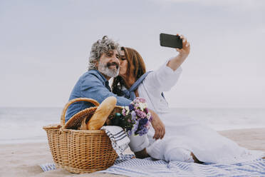Happy senior woman kissing man and taking selfie through smart phone at beach - OIPF03512