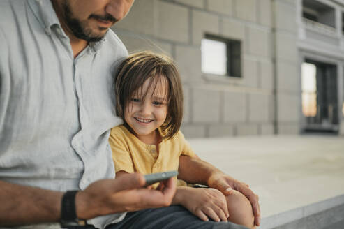 Lächelnder Sohn schaut seinen Vater an, der ein Mobiltelefon benutzt - ANAF02050