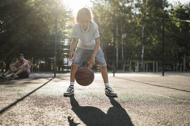 Boy dribbling basketball with father in background - ANAF02058