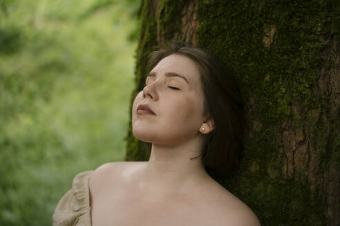 Serene woman leaning on tree in garden - YBF00164