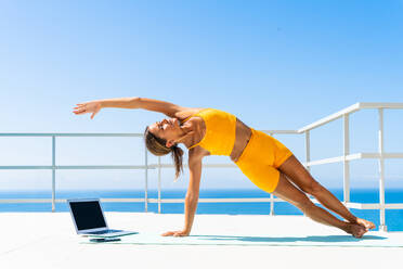 Beautiful young atlethic woman wearing sportswear doing yoga meditation on the terrace rooftop with sea view and blue sky - Yoga practicer training outdoors, healthy lifestyle, sport and meditation concept - DMDF03718