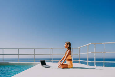 Beautiful young atlethic woman wearing sportswear doing yoga meditation on the terrace rooftop with sea view and blue sky - Yoga practicer training outdoors, healthy lifestyle, sport and meditation concept - DMDF03722