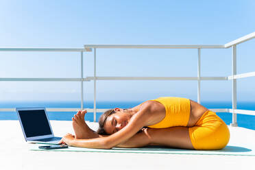 Beautiful young atlethic woman wearing sportswear doing yoga meditation on the terrace rooftop with sea view and blue sky - Yoga practicer training outdoors, healthy lifestyle, sport and meditation concept - DMDF03723